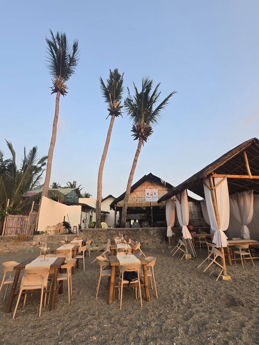 Beachside dining setup at dusk under tall palm trees with the bar hut glowing behind