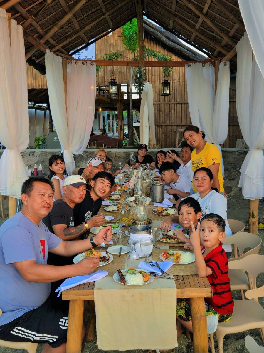 A large family gathered around a long table sharing a meal under the bamboo pavilion