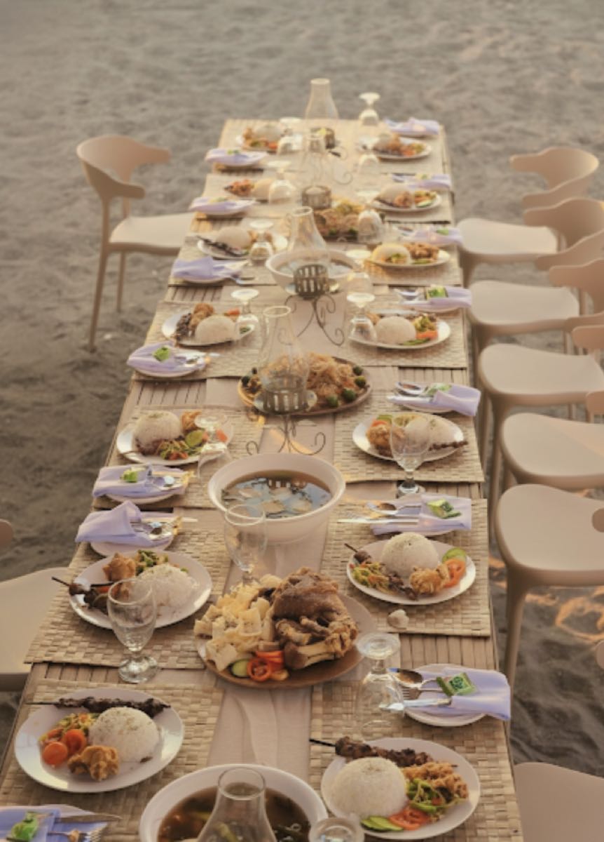 A long communal beachfront dinner table laid out with plates and drinks on the sand at golden hour