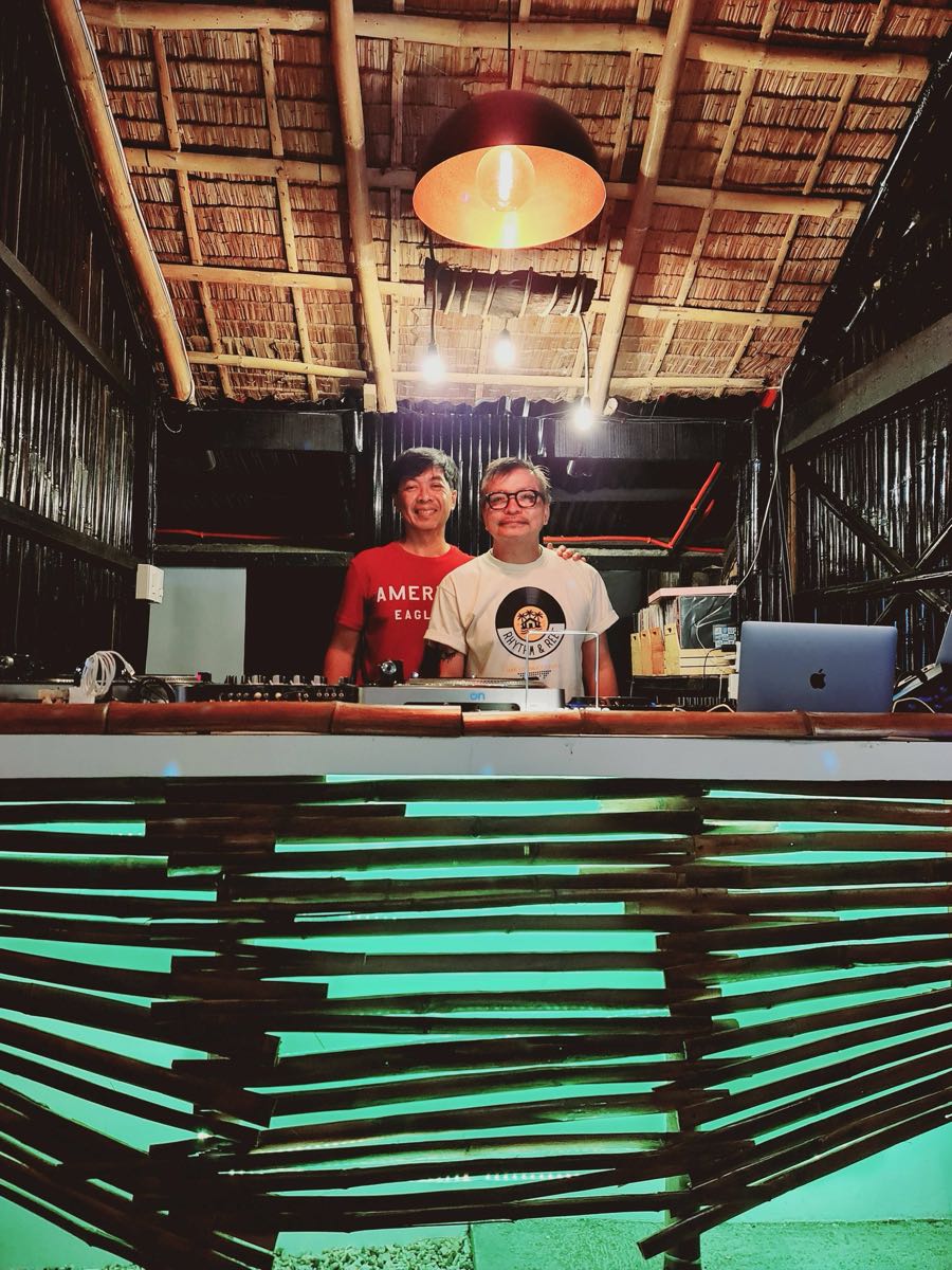 Two DJs behind the oceanfront deck bar at blue hour, with green underlighting and a thatched roof overhead