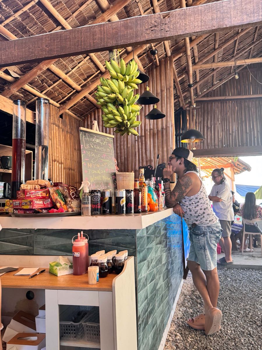 A guest being served at the open-air bar, with hanging bananas and bottles behind the counter